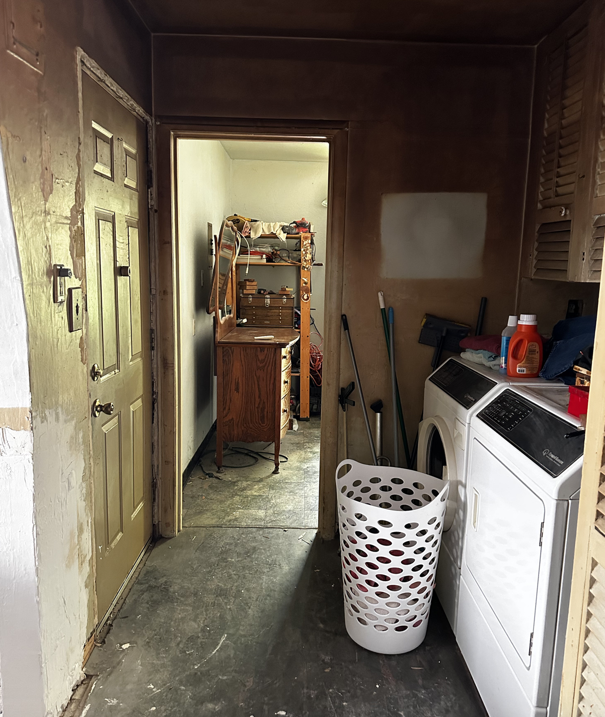 Current laundry and back hallway with peeling paint, bare concrete floor and dated louvered cabinets